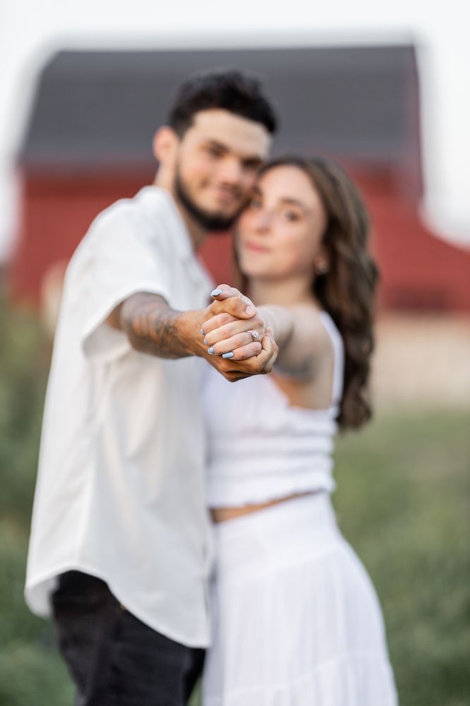 Engaged couple in front of a red barn, holding their joined hands toward the camera with the ring in focus