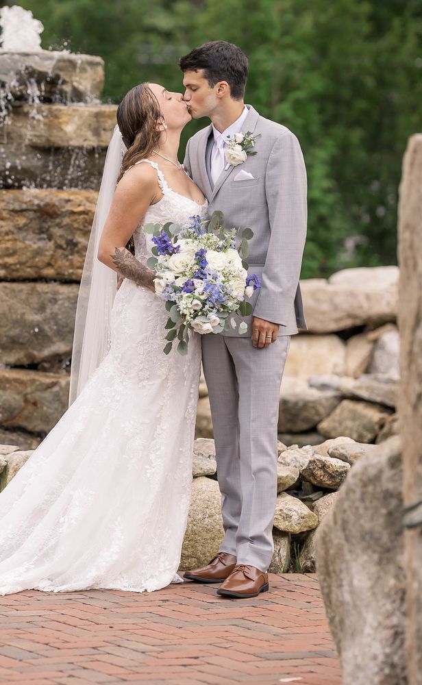 Bride and groom kissing beside a stone fountain, the bride holding a white and blue bouquet